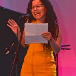 a woman in a yellow dress holding a piece of paper and standing in front of a microphone